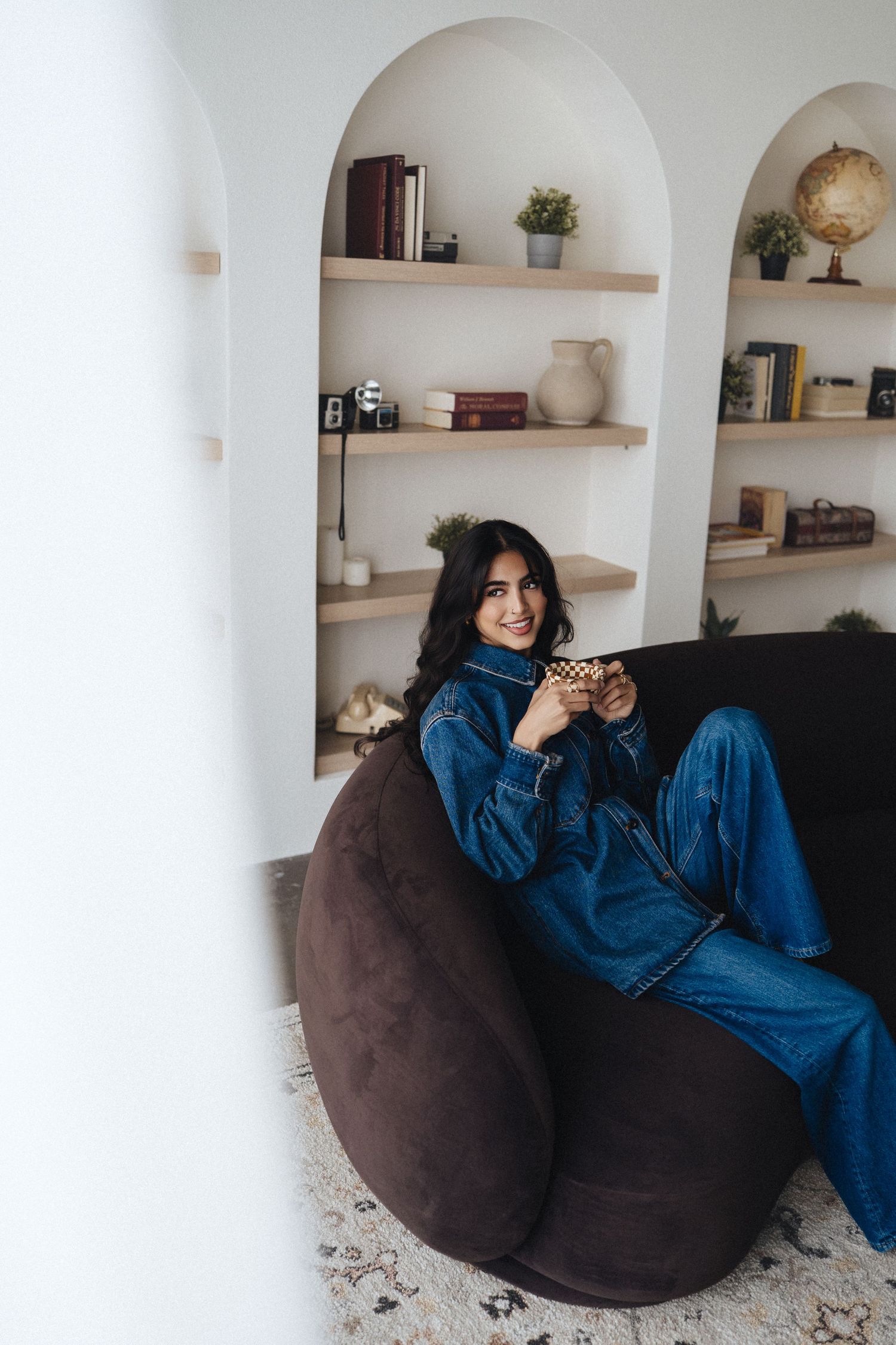 Young woman in denim outfit relaxing on a bean bag chair, smiling and holding a cup in a cozy home interior.