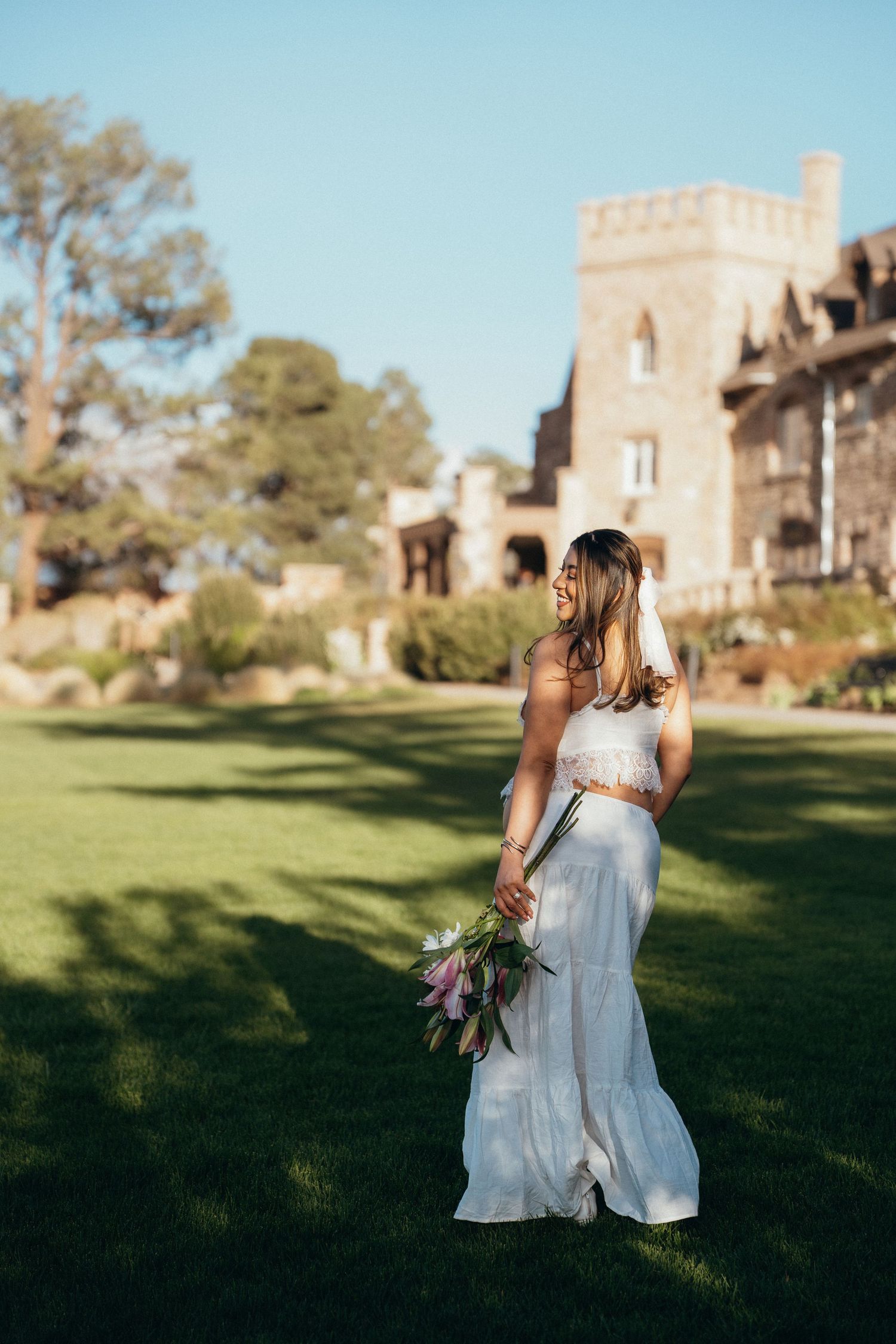 Bride in white two-piece gown holding purple tulip bouquet standing on green lawn before a stone castle.