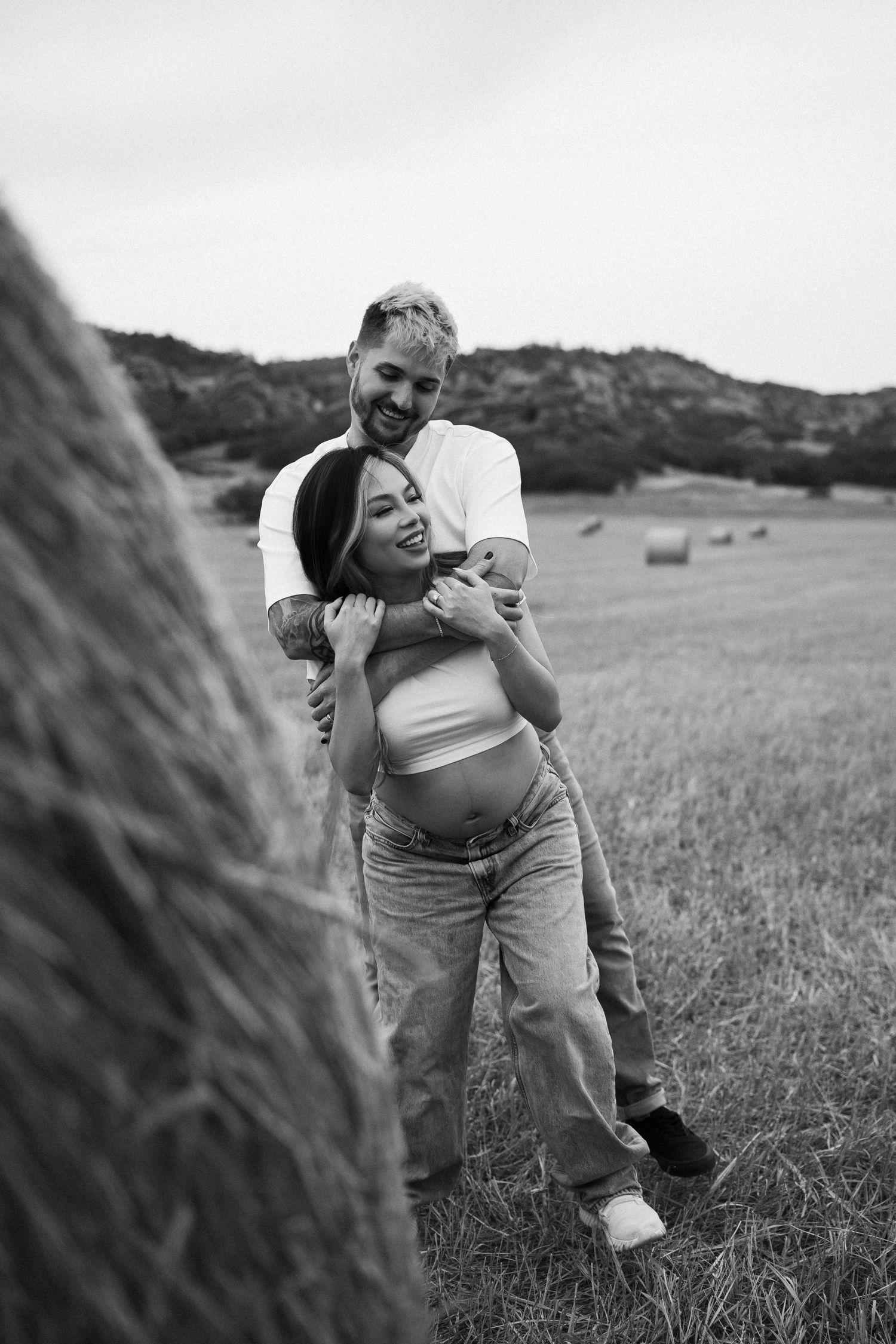 A playful couple embraces and laughs together in a grassy field with hay bales visible in the background.