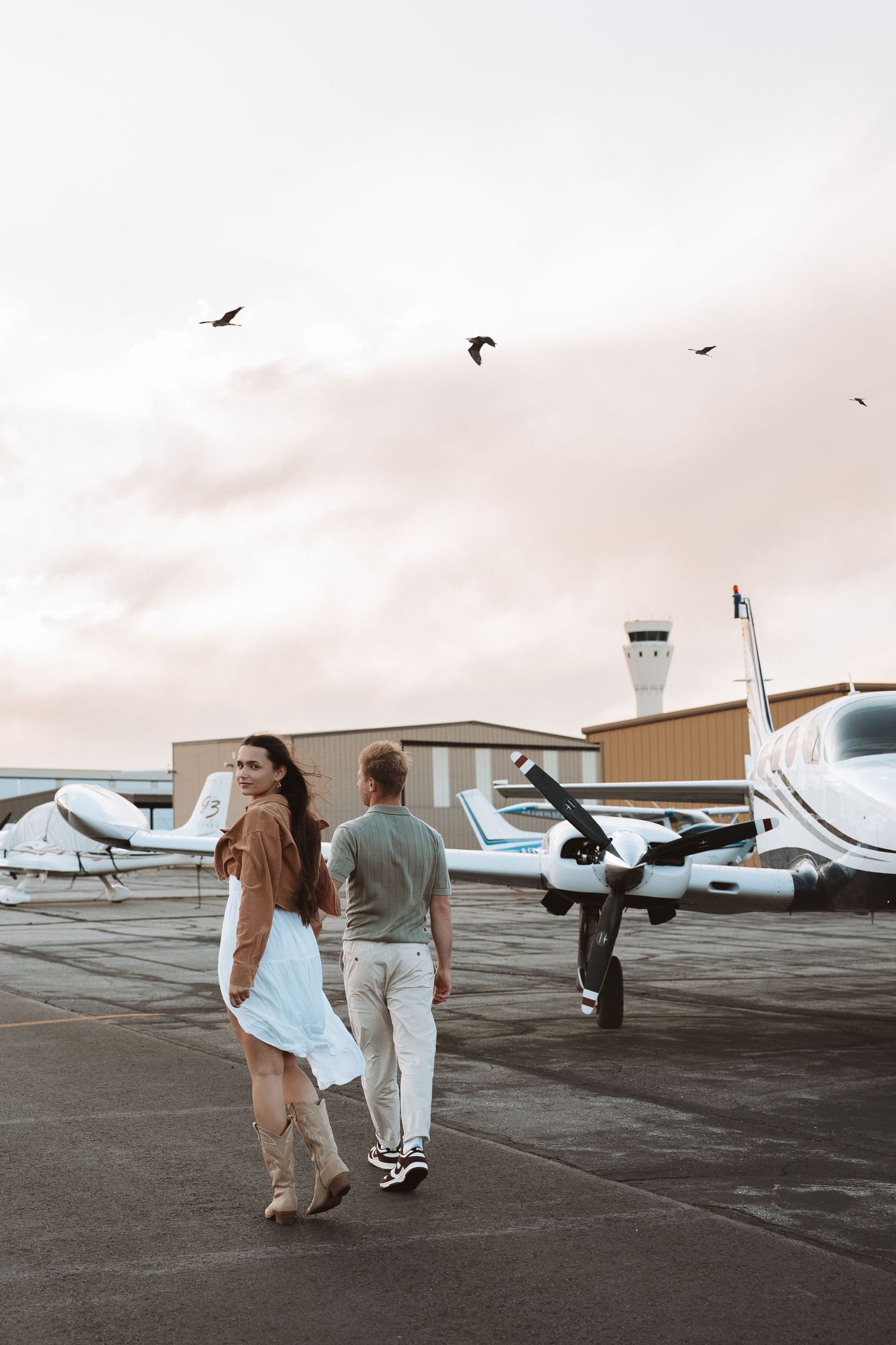 Two people walk across an airport tarmac with small private planes and a control tower visible in overcast conditions.
