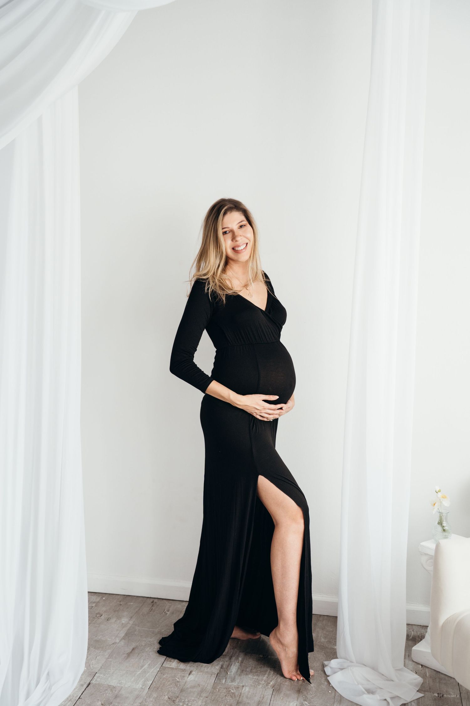 A black floor-length maternity gown with long sleeves and thigh slit stands elegantly against white curtained backdrop.