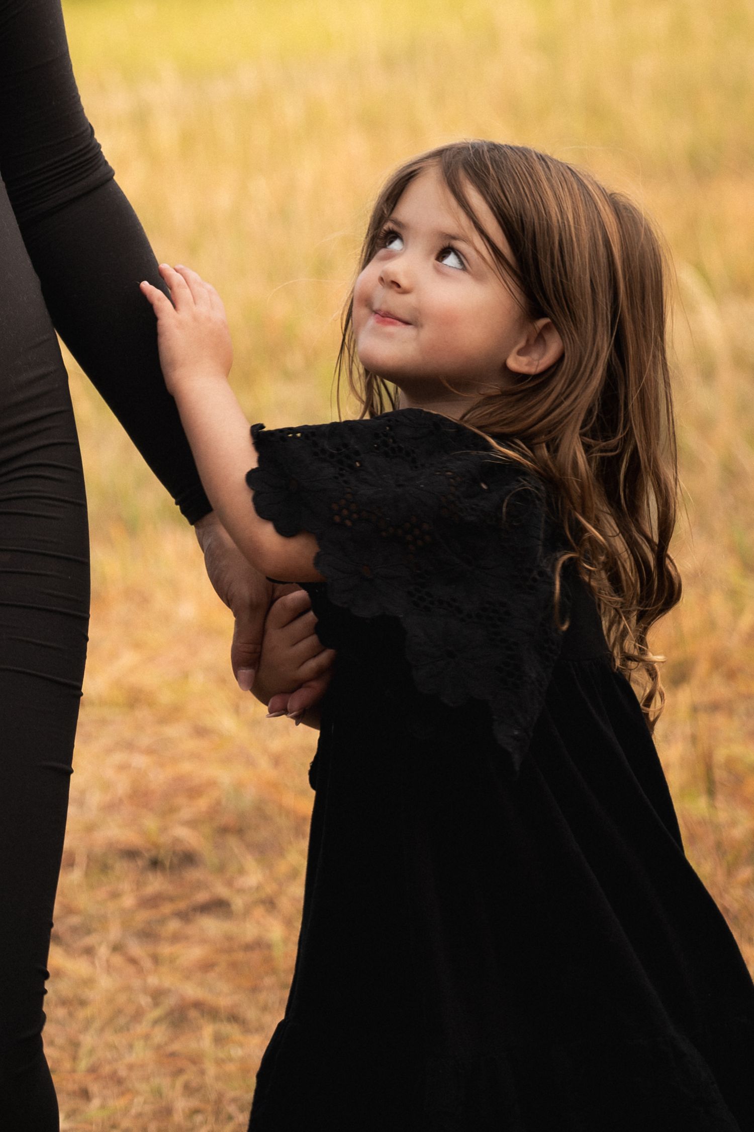 A child in a black dress looks up lovingly while holding onto someone in an outdoor setting.