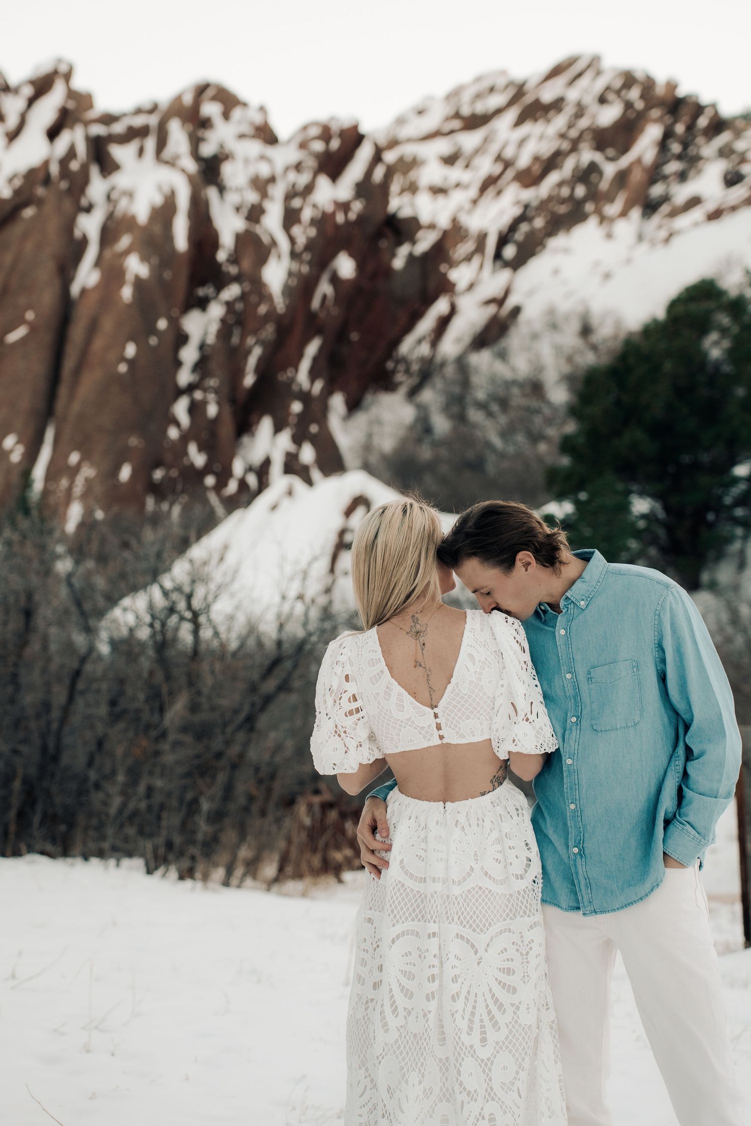 Romantic winter engagement photo of couple in white and blue outfits against snowy mountain backdrop.