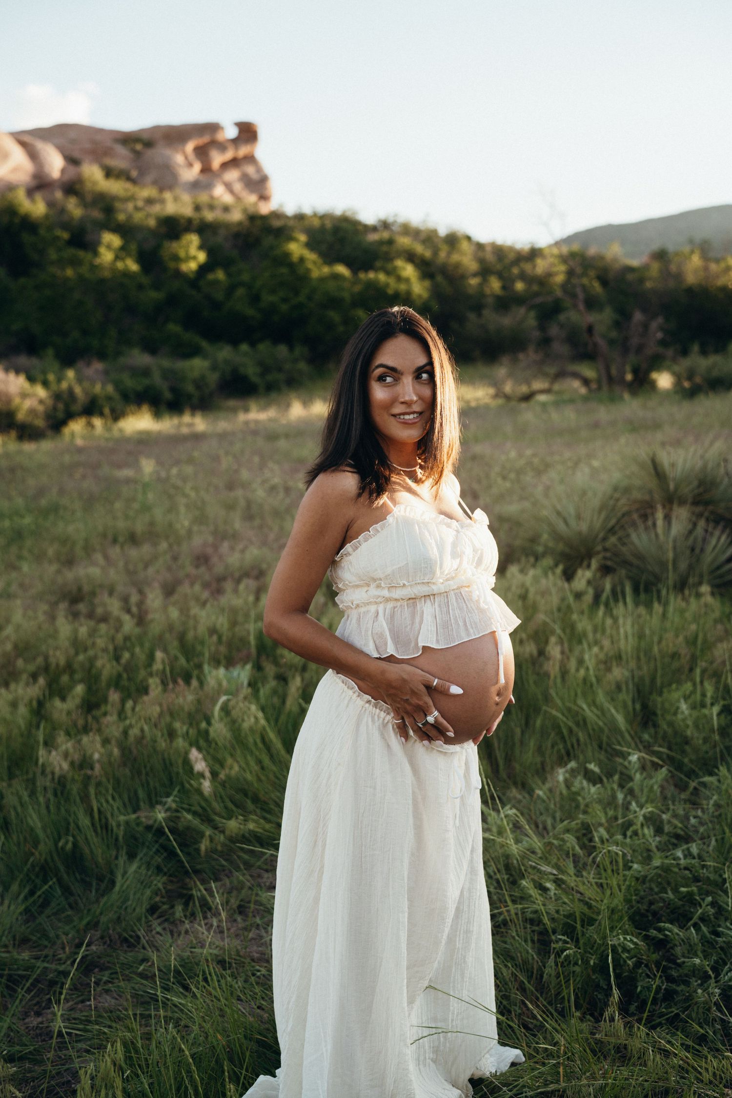 A maternity photo features an expectant mother in a flowing white dress standing in a grassy field at sunset.