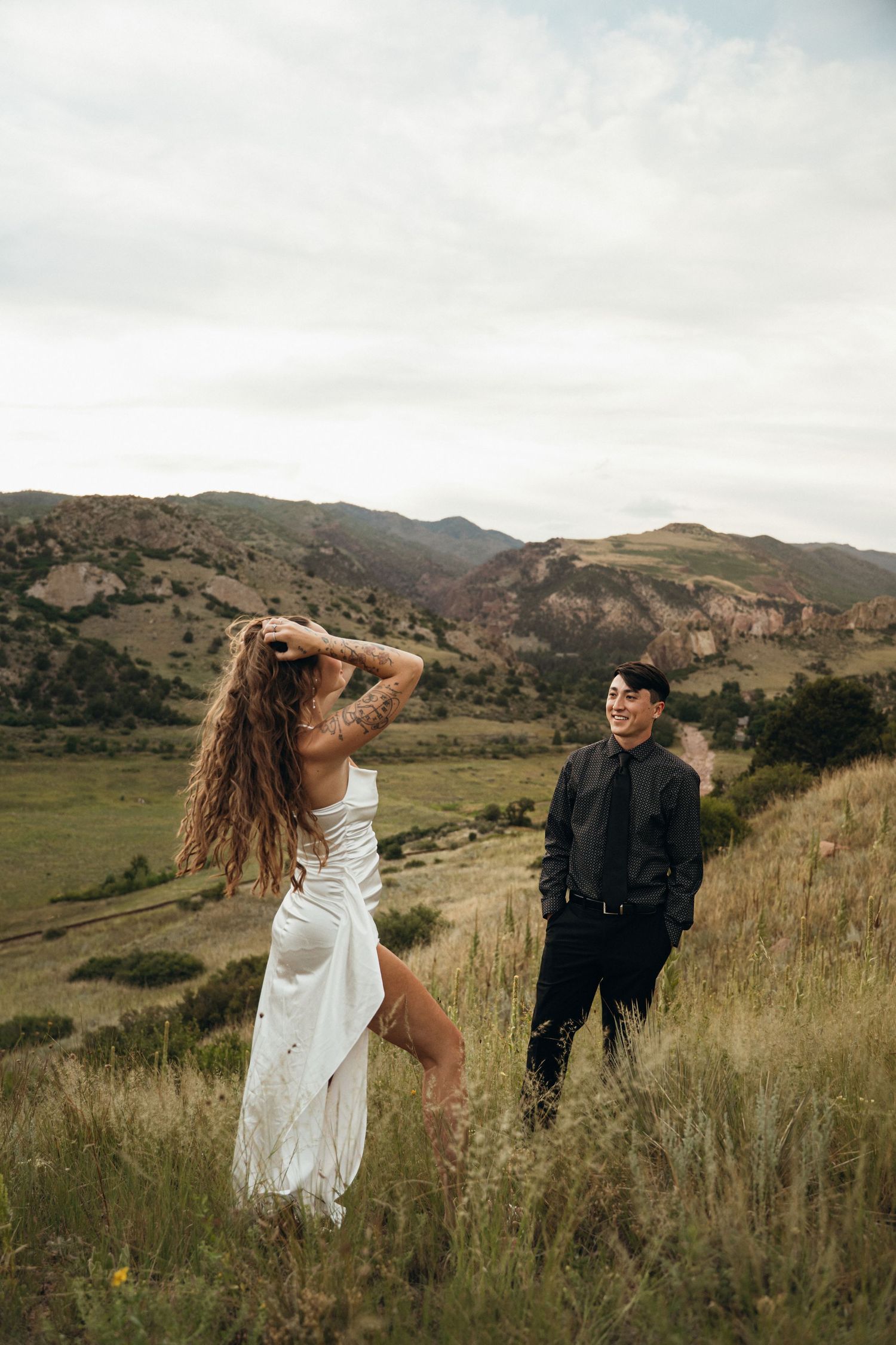A couple in formal wear poses playfully in a scenic mountain meadow with dramatic rock formations behind them.