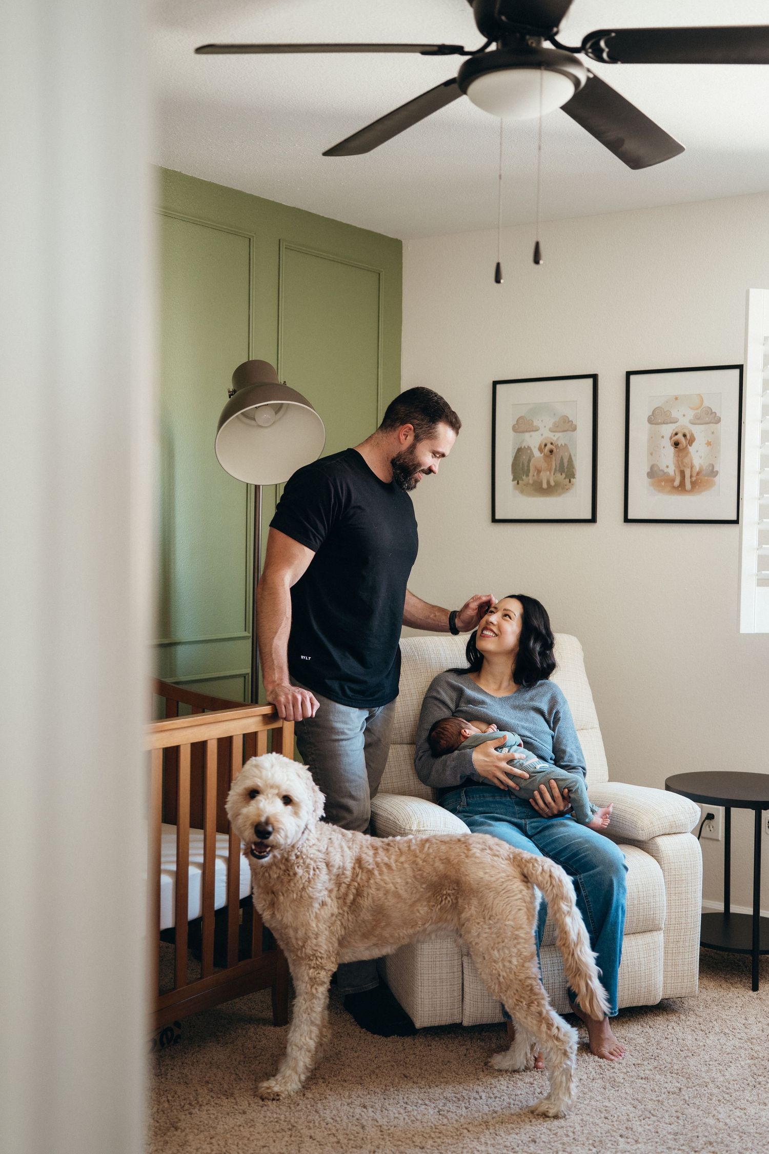 A couple relaxes on a couch with their fluffy white Goldendoodle dog in a modern living room with green accent walls.