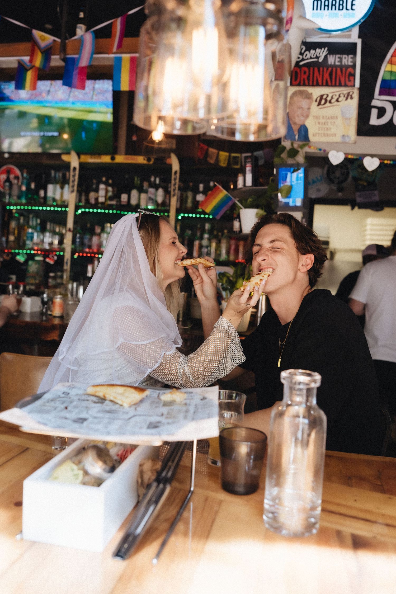 Couple sharing pizza at a bar, bride wearing veil with rainbow pride flag, celebrating at a colorful venue.
