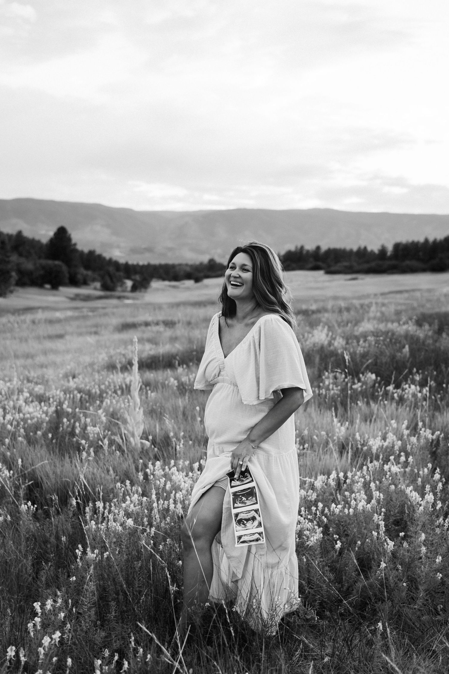 A figure in a white dress poses thoughtfully in a sunlit meadow of wildflowers with mountains in the background.