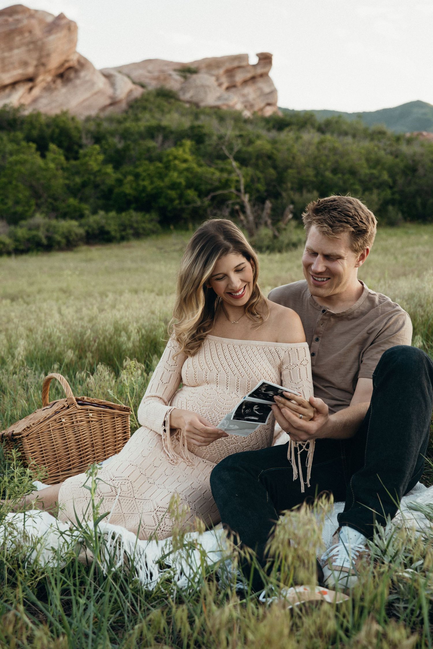 A couple sits on a picnic blanket in a grassy field with red rock formations in the background holding ultrasound photos.