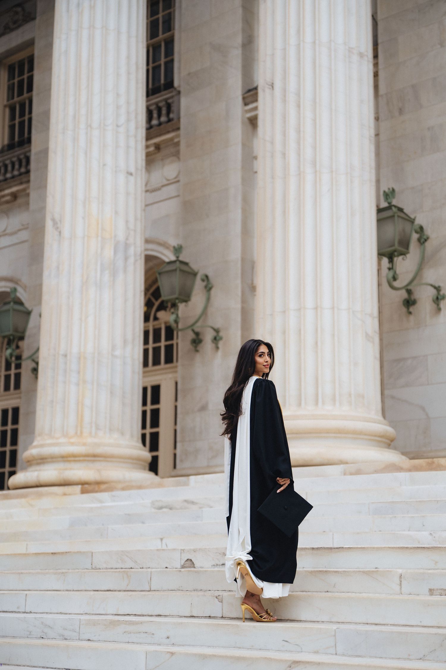Graduate in black gown poses on marble steps before grand classical columns of prestigious university building.
