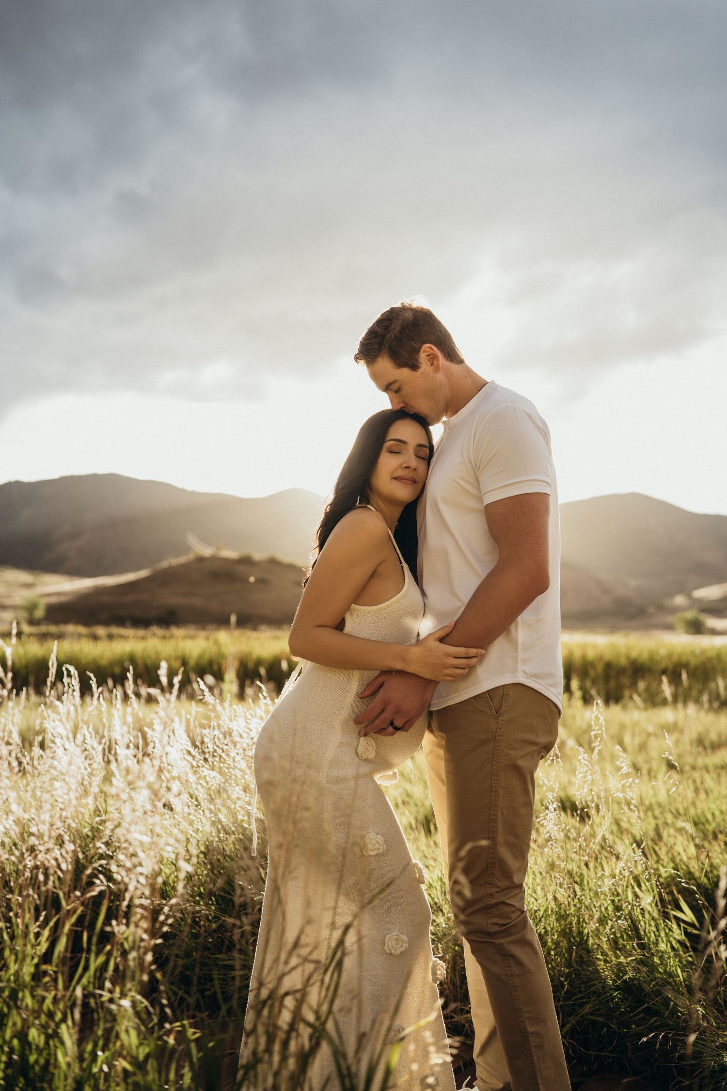 A romantic couple embraces in a sunlit meadow with tall grass and mountains in the background during golden hour.