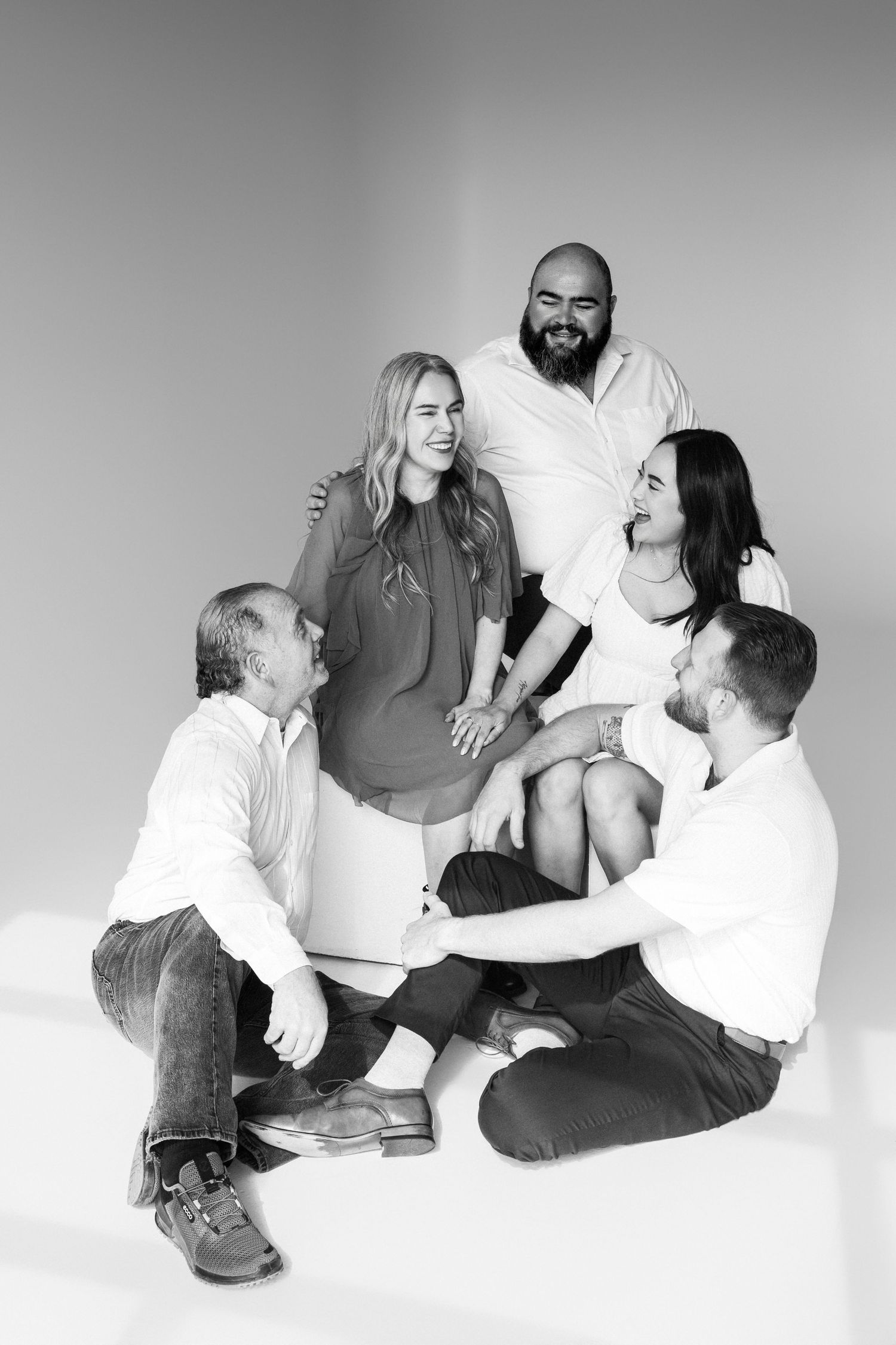 A black and white studio portrait of a group of people laughing and sitting together on the floor.
