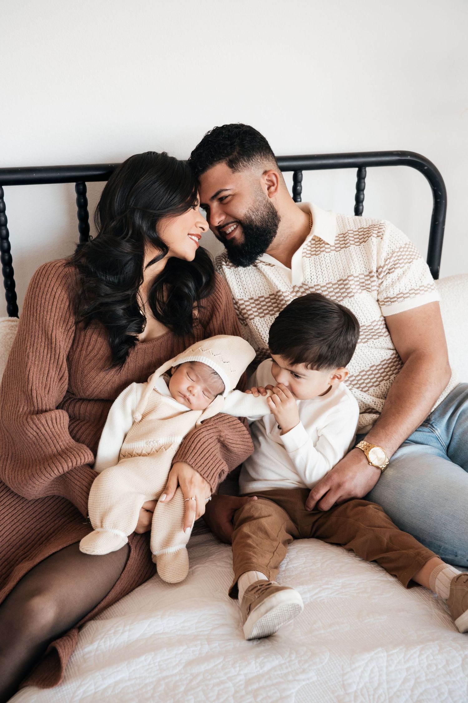 A loving family relaxes together on a bed, sharing a tender moment in matching neutral-toned outfits.
