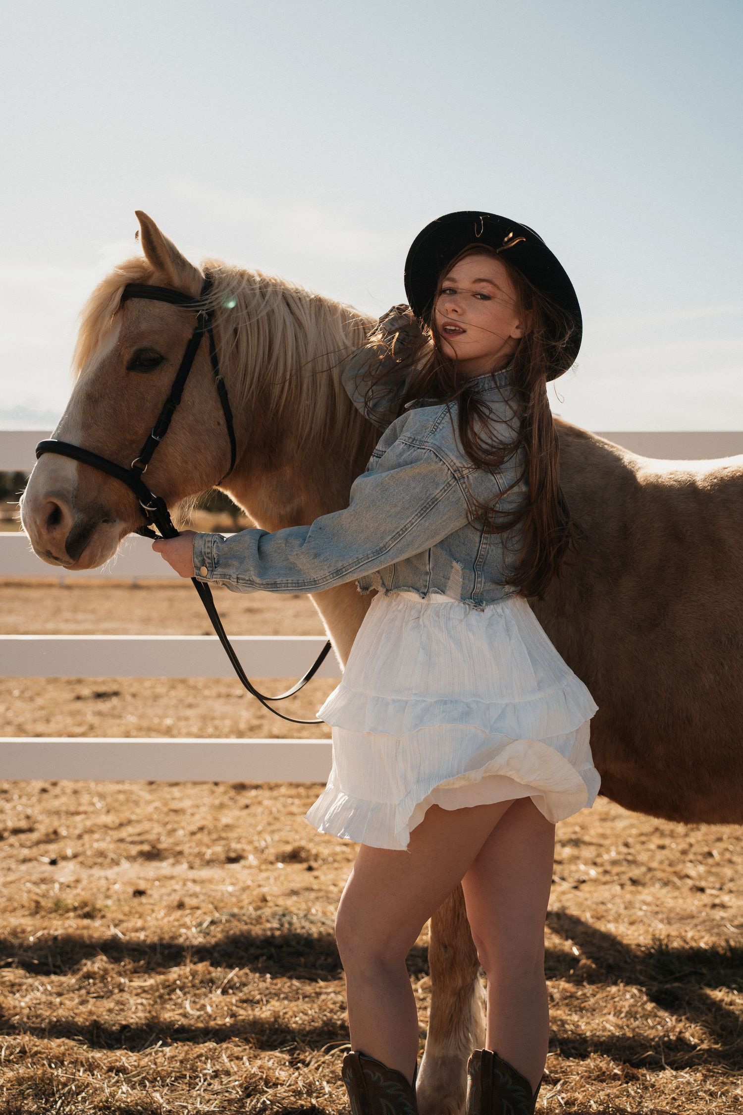 A brown horse stands with someone in a denim jacket, white skirt and black hat in a sunny field.