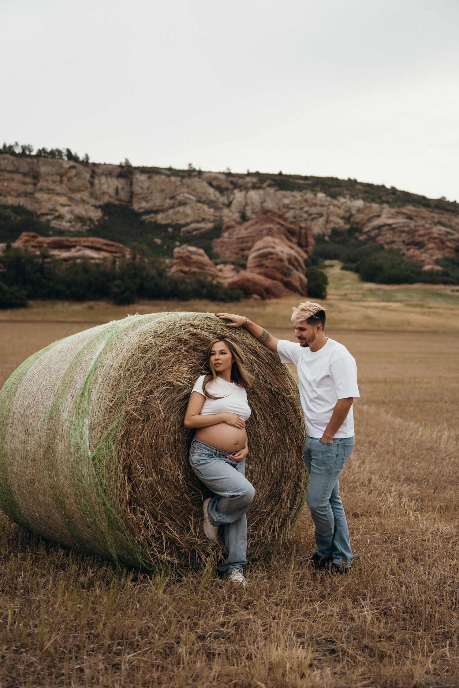 A couple poses by a large hay bale in a field with rocky hills in the background during a maternity photoshoot.