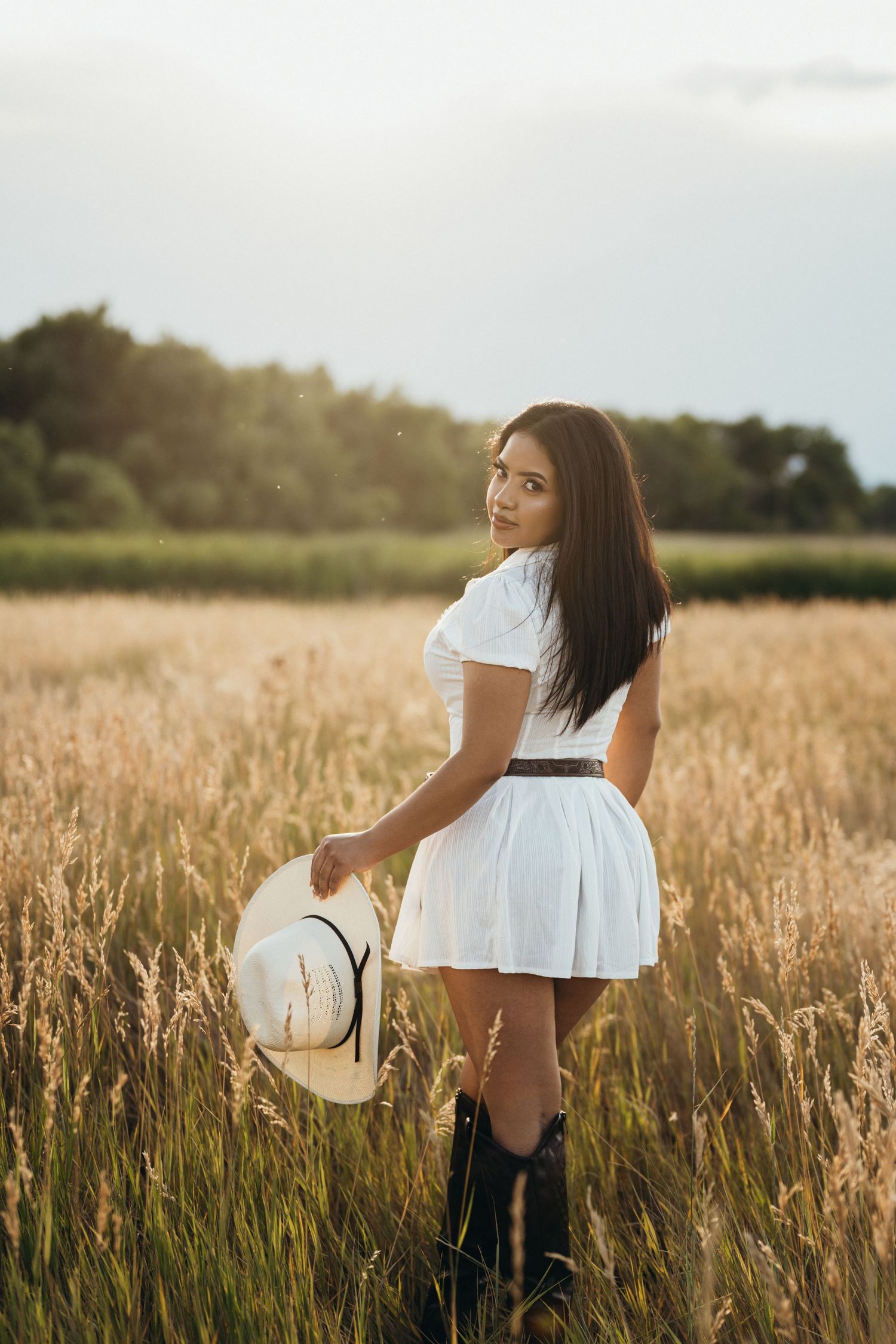 A person in a white dress and cowboy boots stands in a golden wheat field at sunset holding a straw hat.