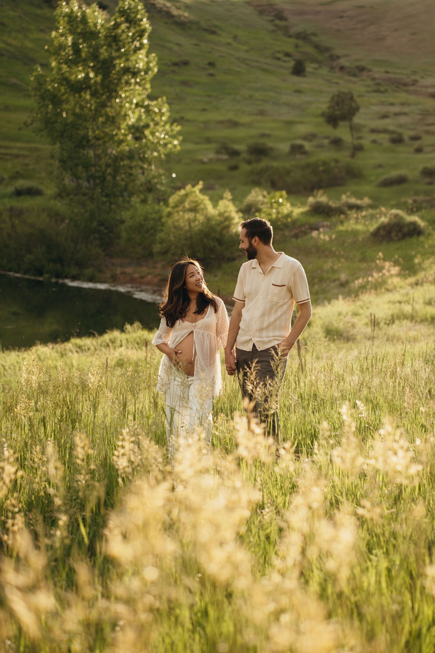 Couple walking together through a sunlit meadow with golden grass and rolling hills in the background during sunset.