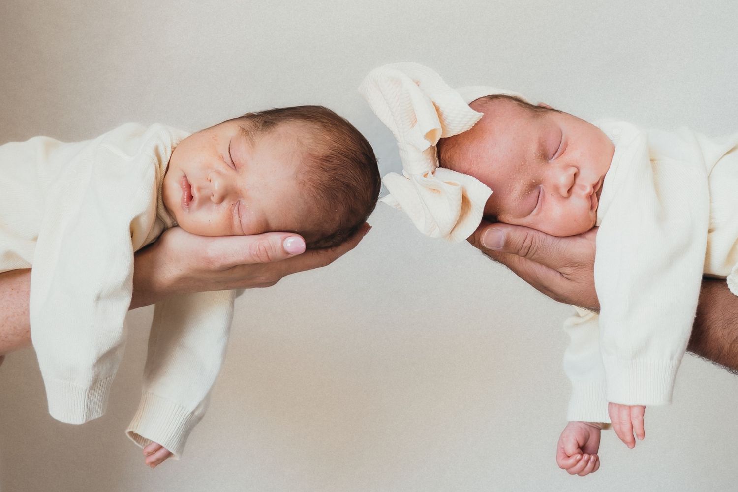 Two newborn babies swaddled in white blankets rest peacefully while being gently cradled.