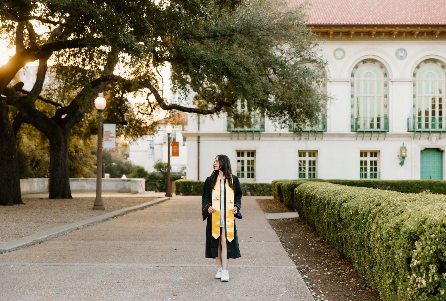 Graduate in black cap and gown with gold stole walks campus path at sunset.