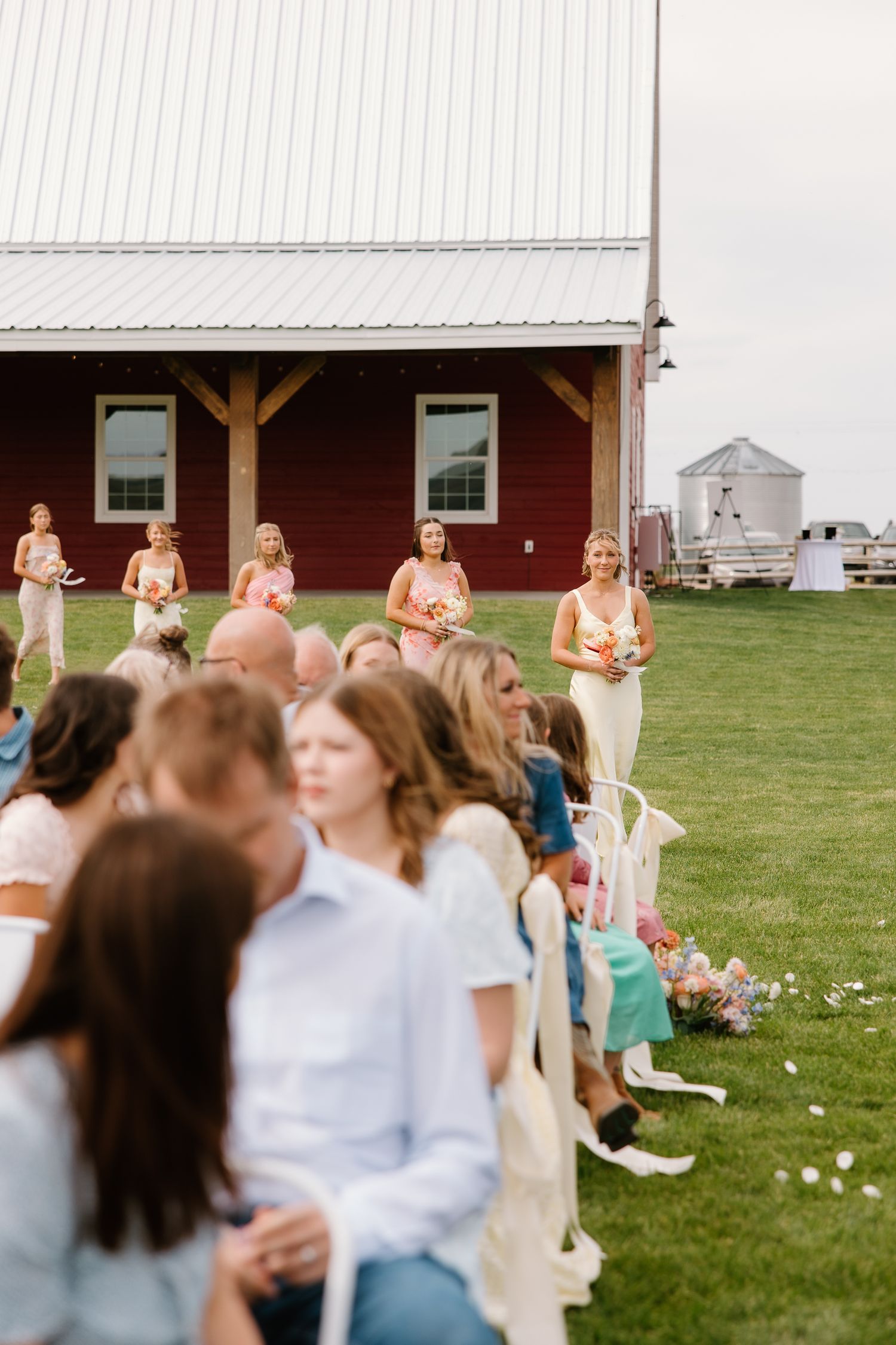 Bridesmaids in cream dresses walk down the aisle at an outdoor barn wedding.