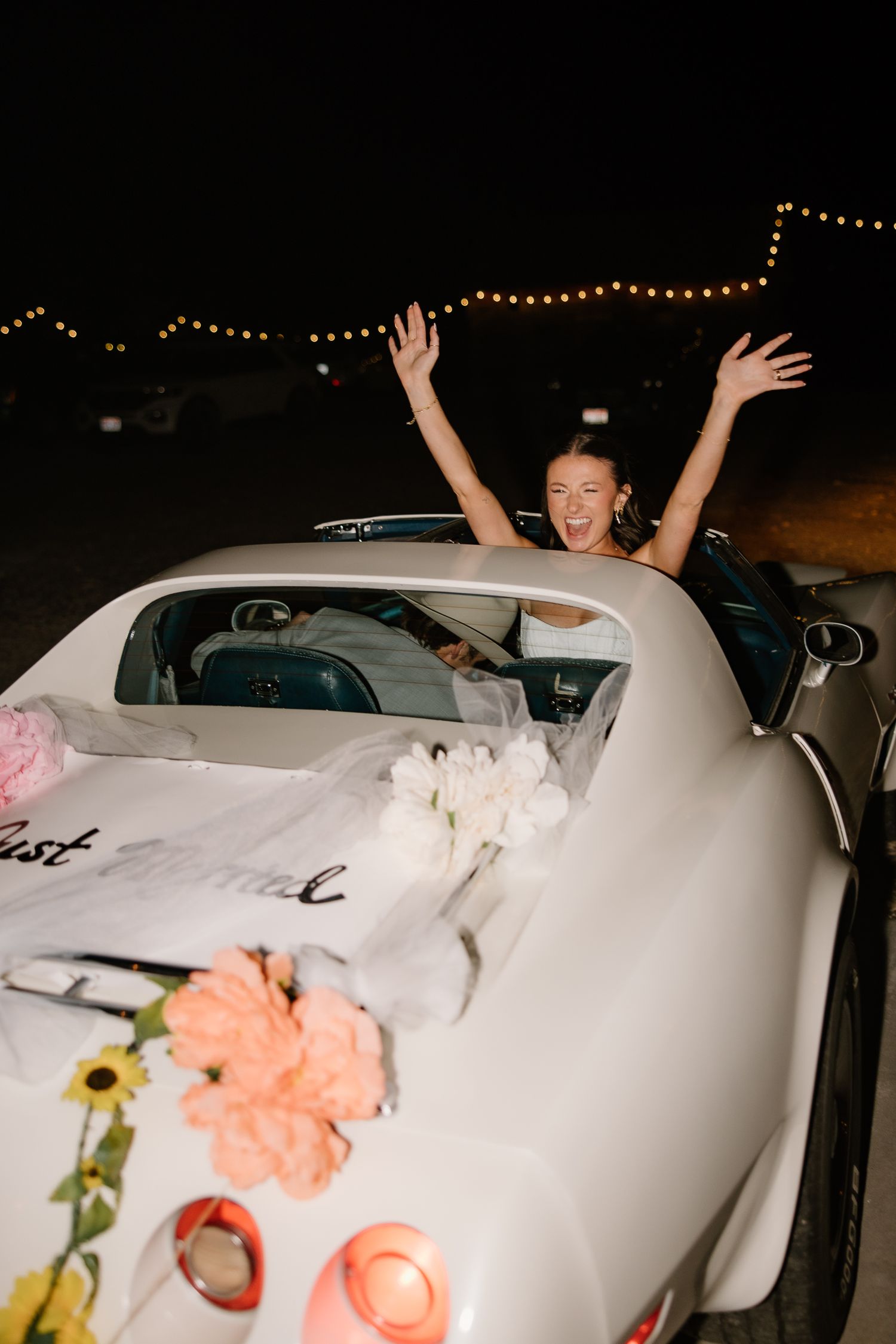 Joyful bride raises arms in vintage white convertible decorated with flowers after wedding.