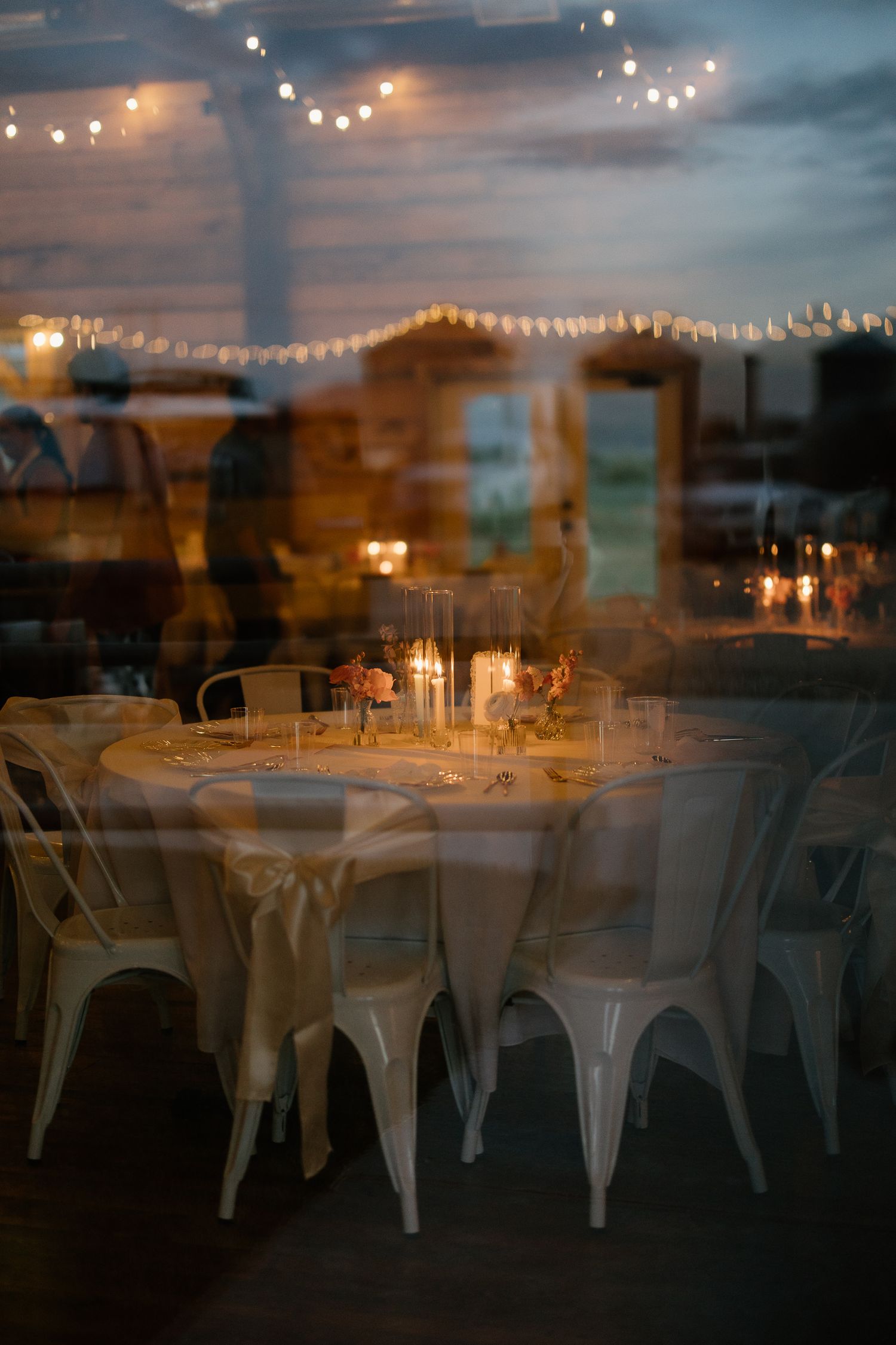 Elegant candlelit wedding reception tables with string lights reflected through glass at dusk.