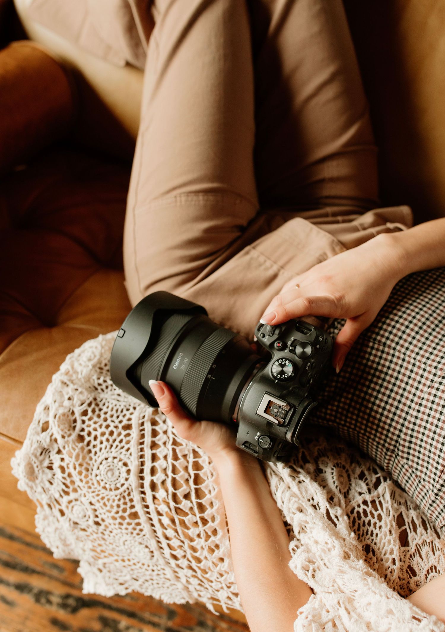 A person sitting on a leather sofa holding a black DSLR camera with a wide lens over a lace cloth.
