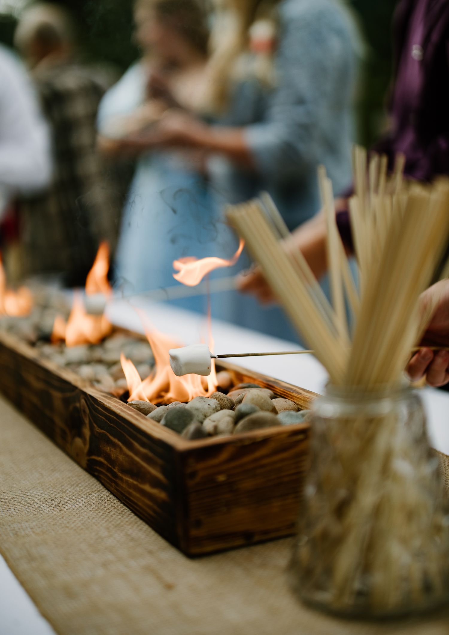 Rustic wooden fire centerpiece with river rocks and bamboo skewers at outdoor wedding reception.