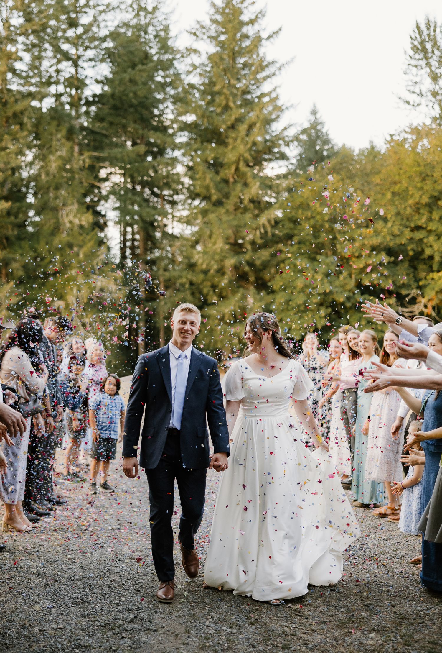 Newlyweds walking through confetti shower with cheering guests lining path among tall evergreen trees.