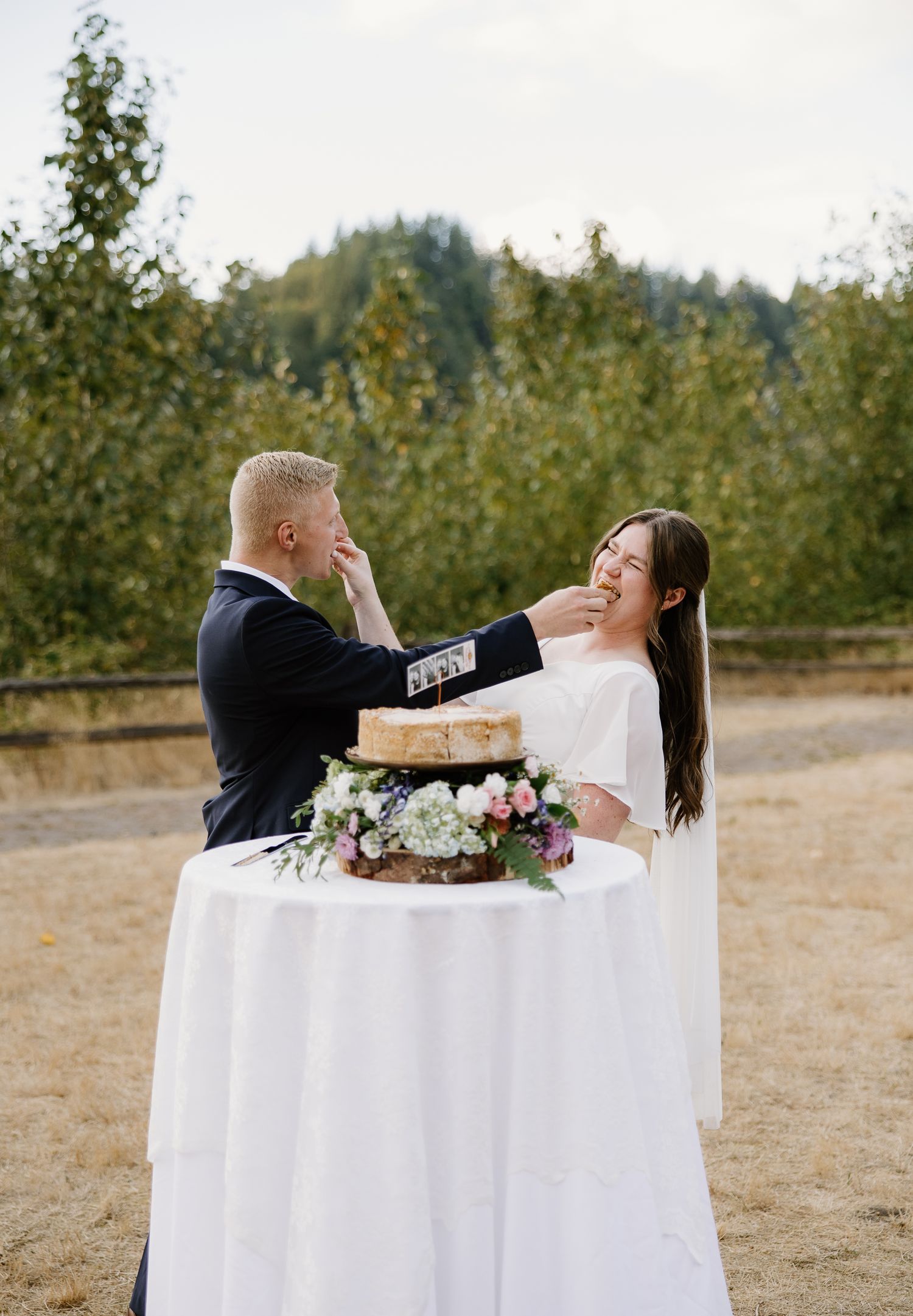 Bride and groom laugh while feeding each other wedding cake at an outdoor reception.