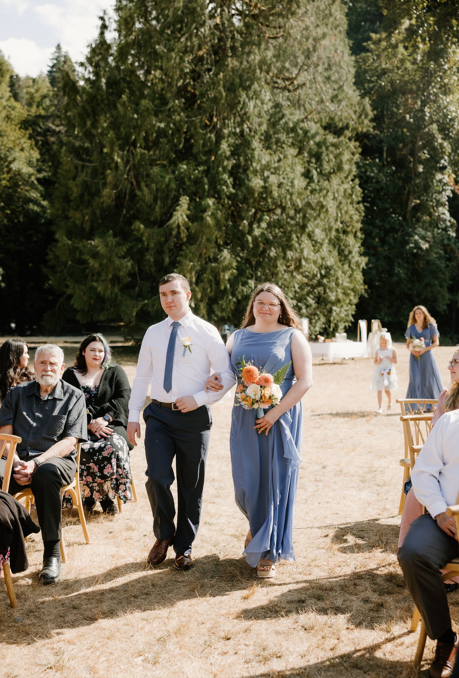 Bridesmaid in blue dress and groomsman walk down outdoor wedding aisle carrying colorful floral bouquet.