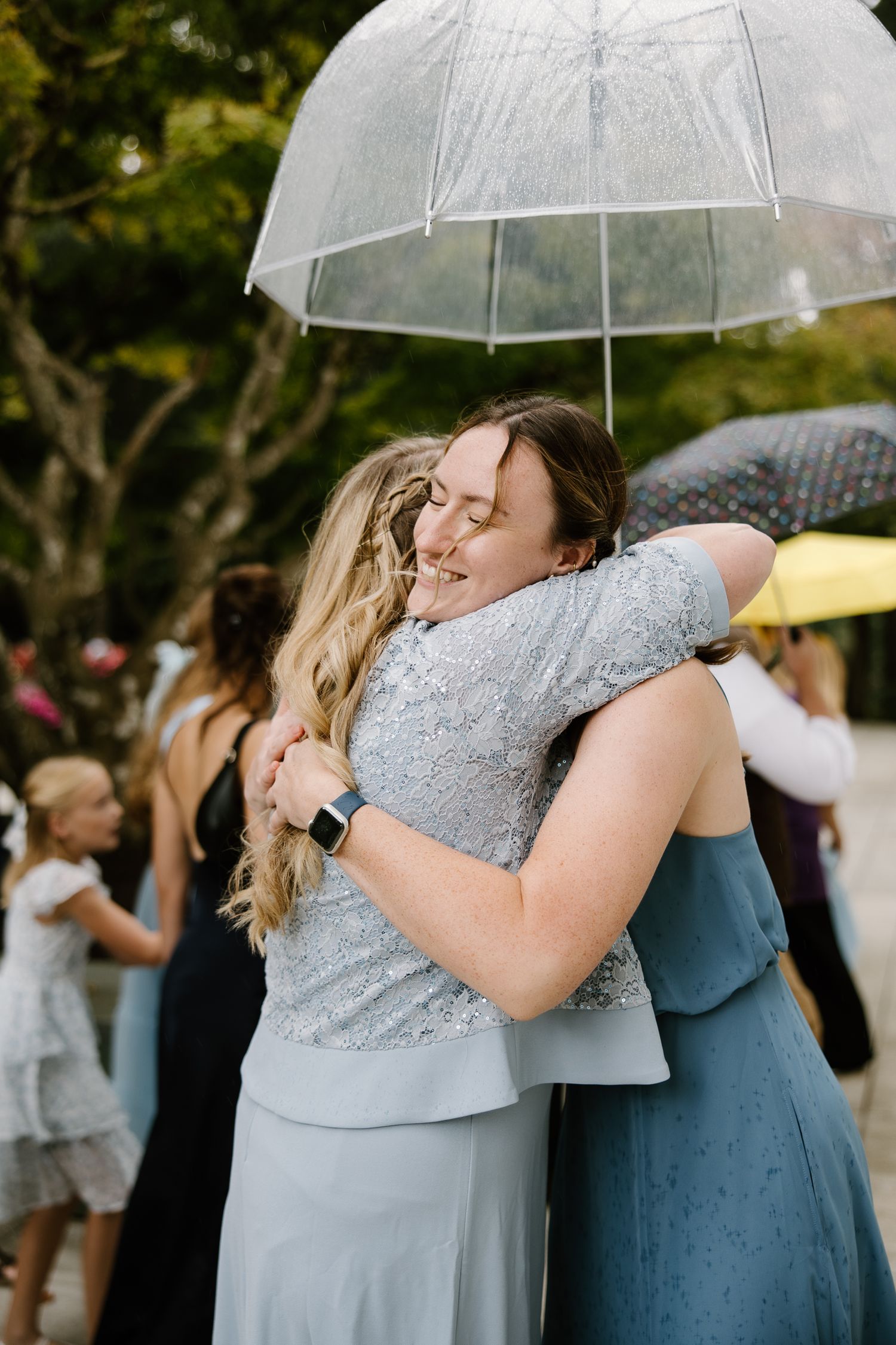 Two women in blue dresses embrace warmly under a clear umbrella at an outdoor wedding event.