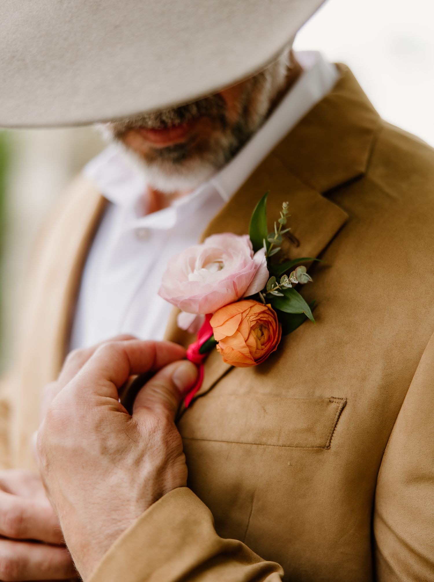 Groom in tan suede jacket and cowboy hat wearing a pink and orange ranunculus boutonniere at a wedding.