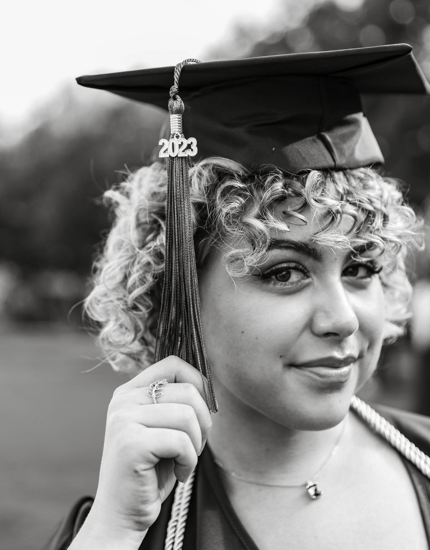 Graduate in cap and gown holds tassel with 2023 charm in black and white photo.