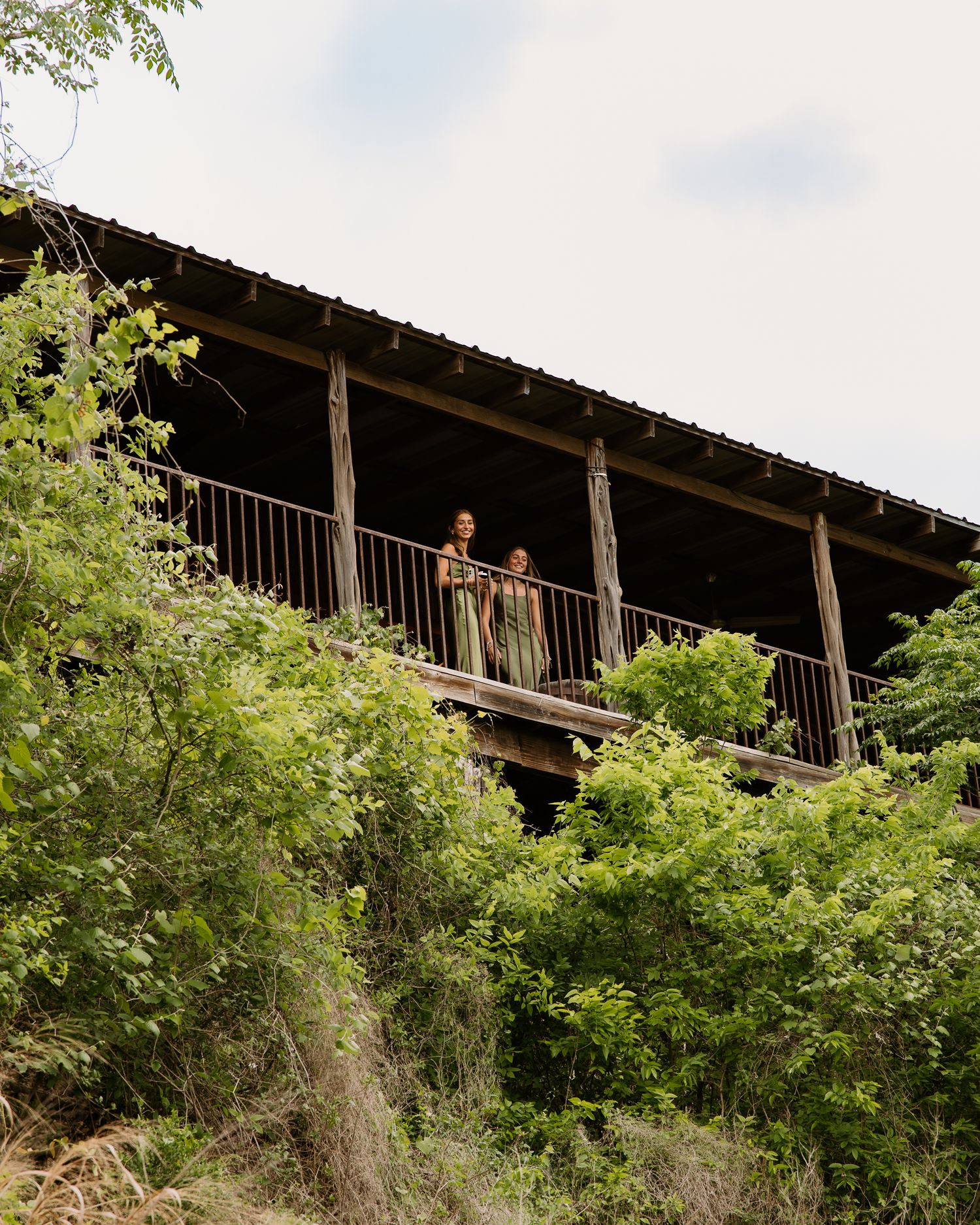 Two women in green dresses stand on rustic wooden balcony overlooking lush green hillside at wedding venue.