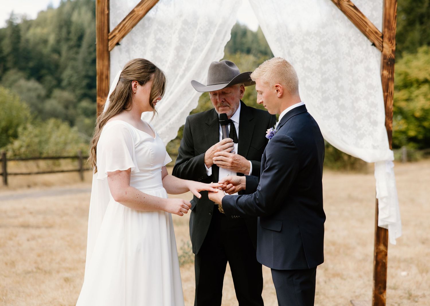 Bride and groom exchange rings at rustic wooden arch altar with white draping during outdoor ceremony.