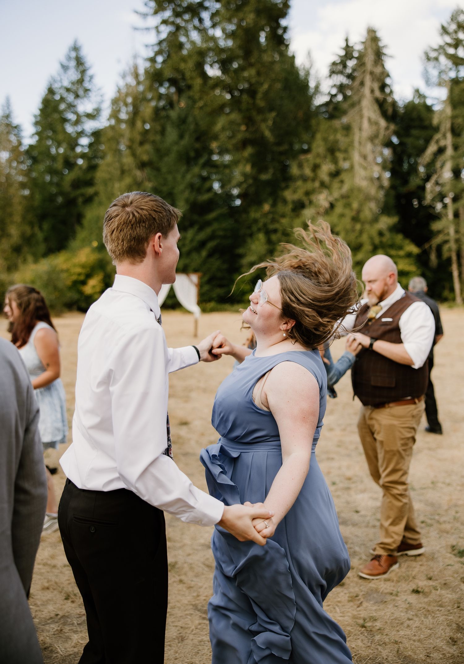 Guests dance energetically at outdoor wedding reception surrounded by tall evergreen trees.
