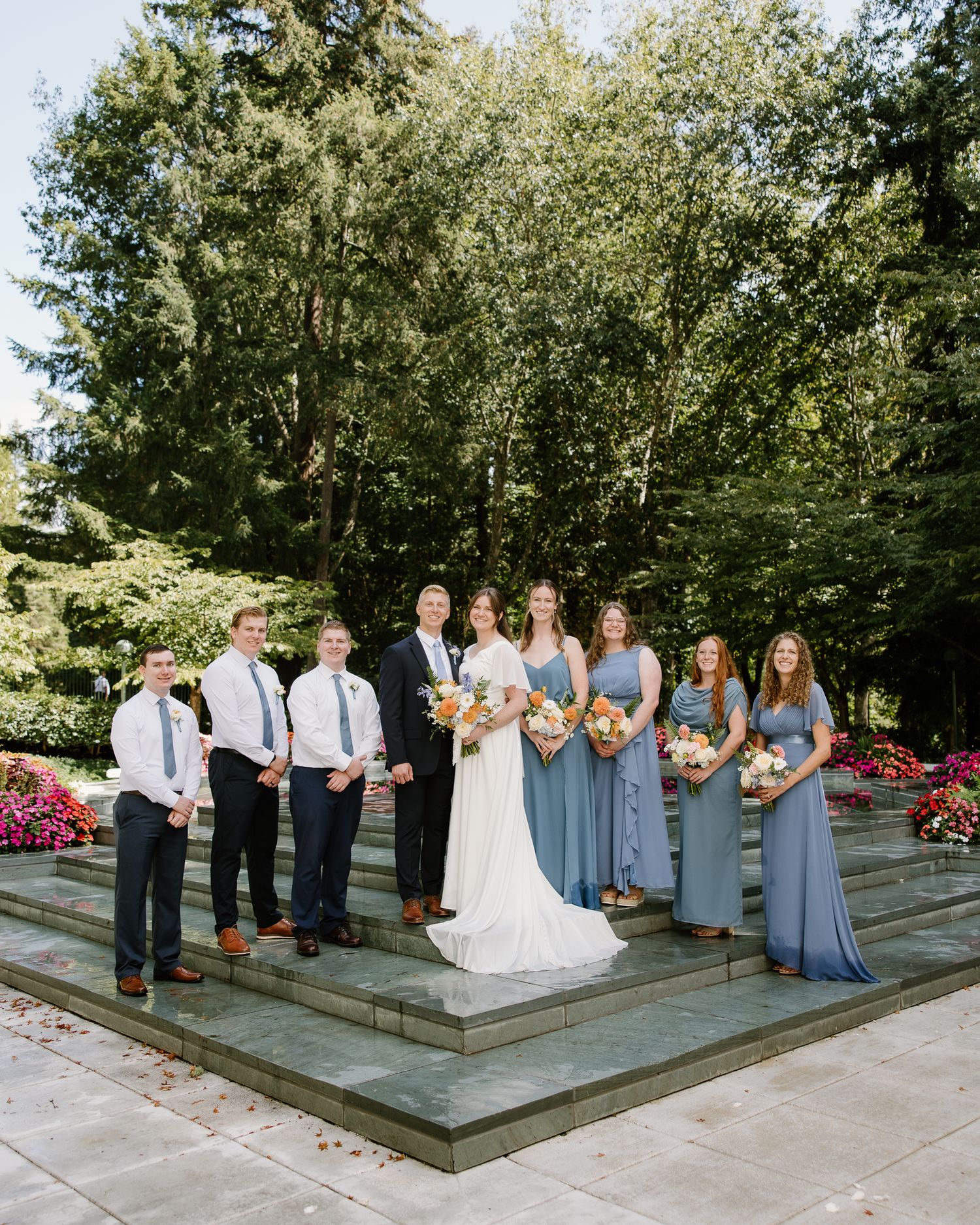 Wedding party poses on stone steps, bridesmaids in blue dresses and groomsmen in navy suits flanking the bride and groom.