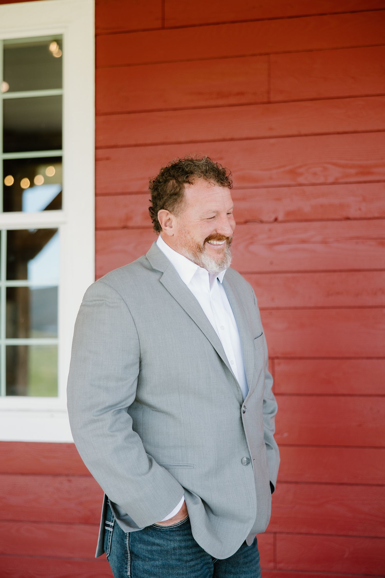 Smiling man with curly hair and beard wearing a gray blazer standing outside a red barn wedding venue.