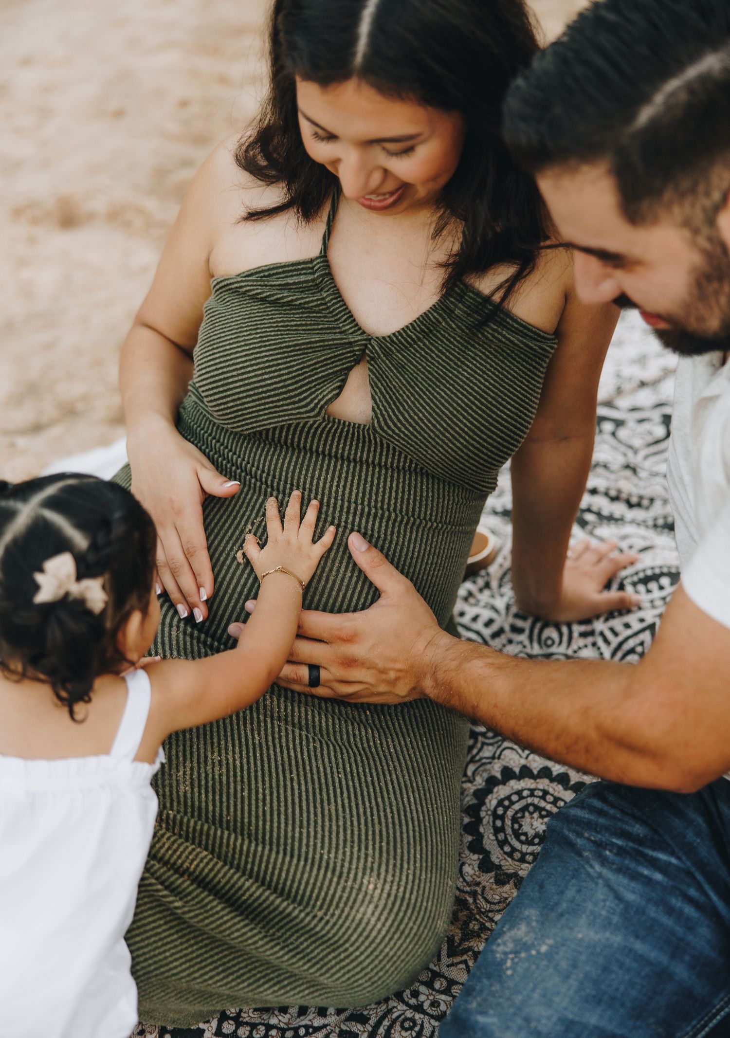 Pregnant woman in green dress with husband and toddler daughter touching her baby bump outdoors.