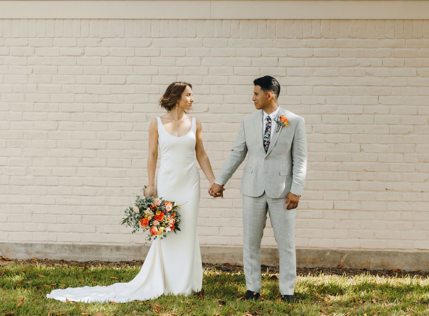 Bride in white gown and groom in light gray suit hold hands while smiling at each other against a white brick wall.