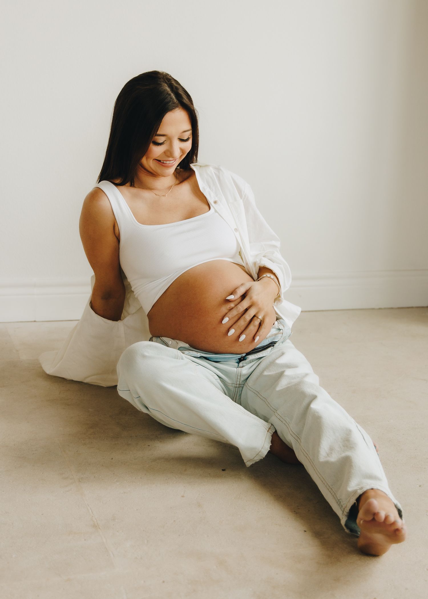 Pregnant woman in white crop top and jeans sits on floor cradling baby bump during maternity photoshoot.