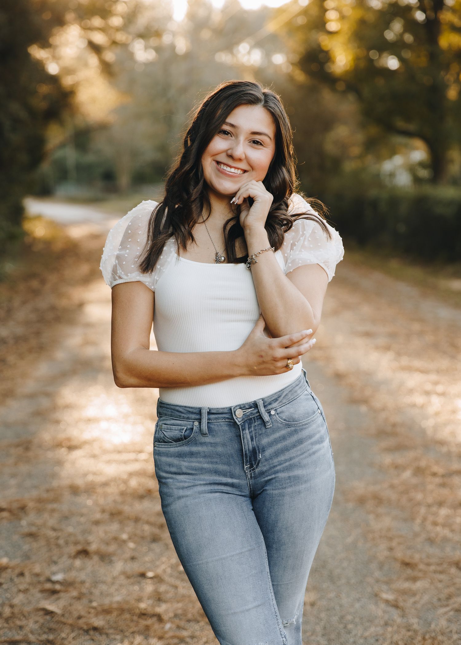 Smiling brunette woman in white top and jeans posing on a sunlit dirt path surrounded by trees.