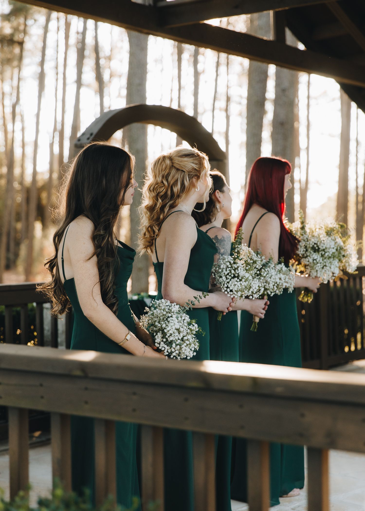 Three bridesmaids in emerald green dresses hold baby's breath bouquets under wooden gazebo at outdoor wedding.
