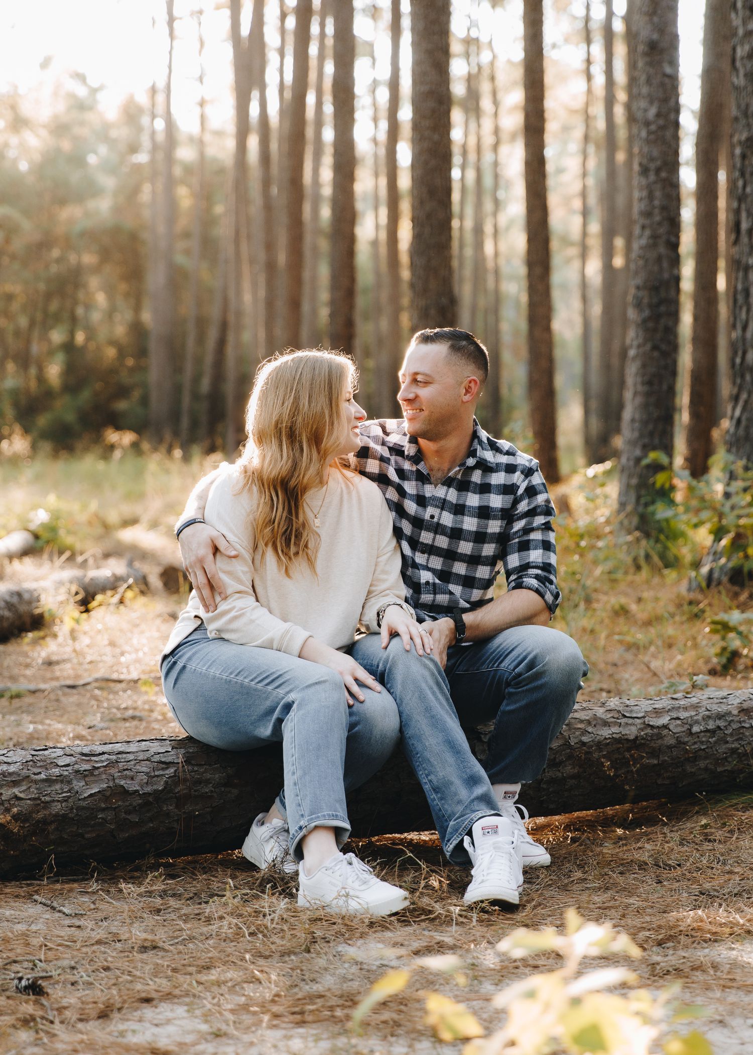 A smiling couple sits together on a fallen log in a sunlit pine forest during a portrait session.