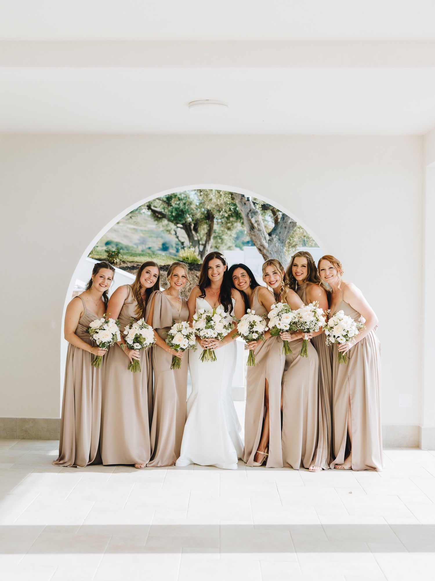 Bride with bridesmaids in champagne gowns holding white floral bouquets, posing under an arched backdrop outdoors.