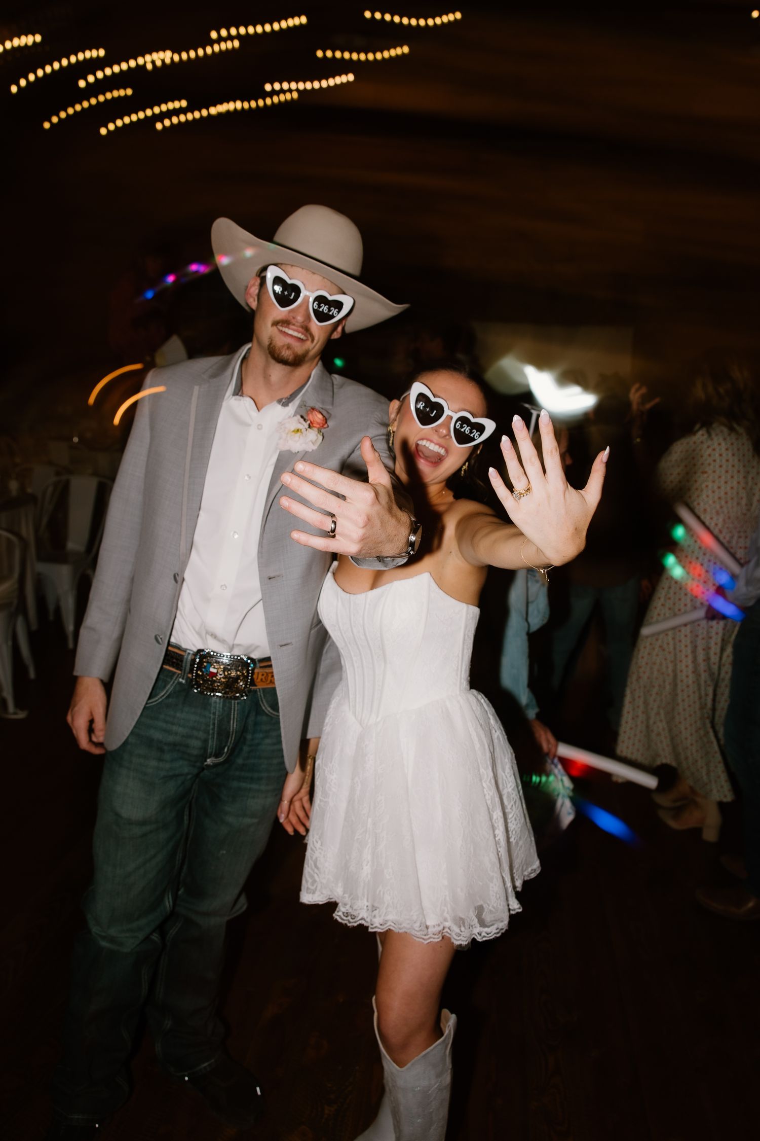 Wedding couple in matching heart sunglasses showing rings on dance floor, groom in cowboy hat.