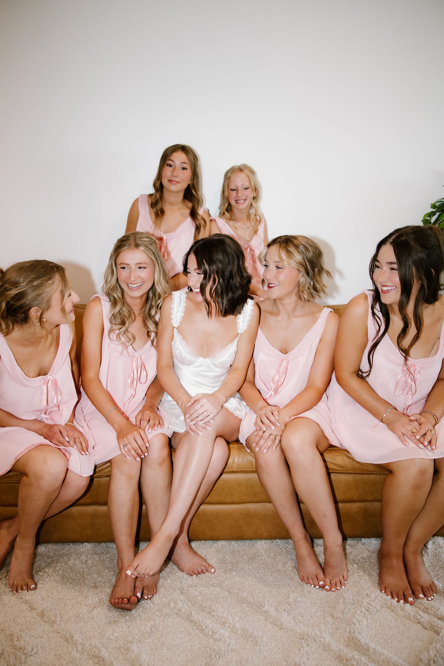 Bride laughing with bridesmaids in matching pink dresses seated together on a couch during wedding preparations.
