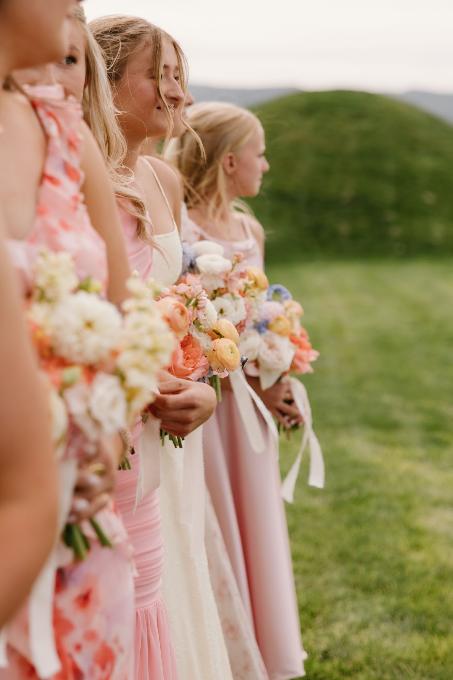 Bridesmaids in pink dresses holding colorful floral bouquets lined up outdoors on a green hillside.