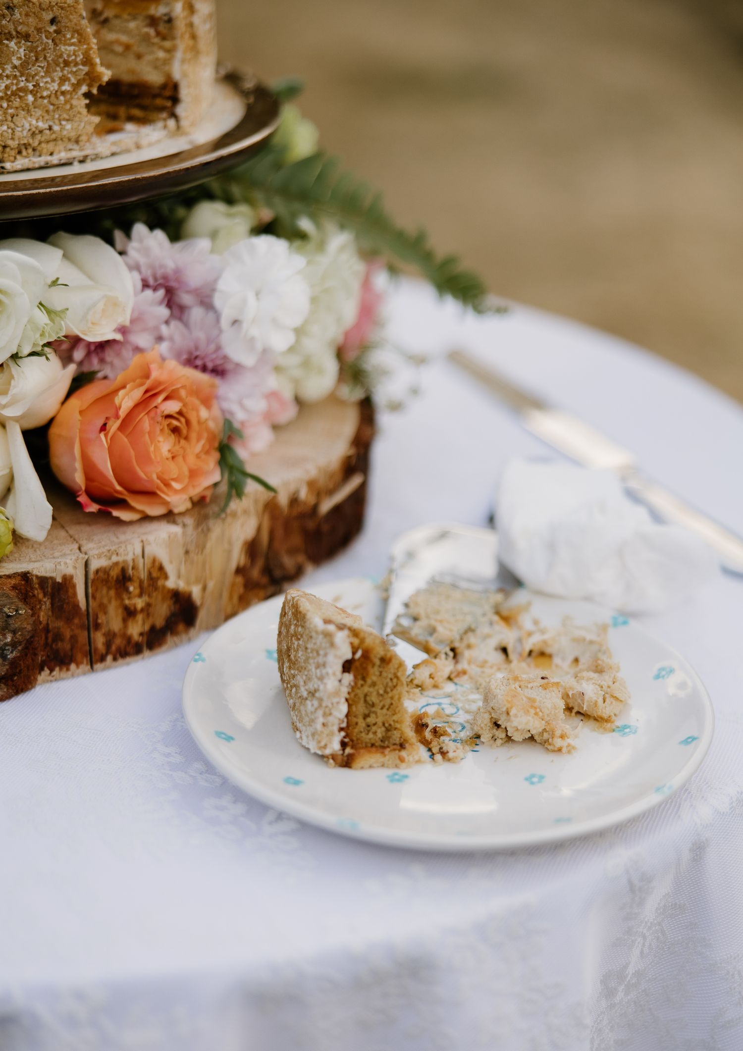 Sliced rustic naked wedding cake on wood slab stand adorned with fresh peach and white flowers.